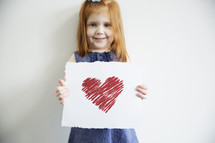 A little girl holds a drawing of a red heart.