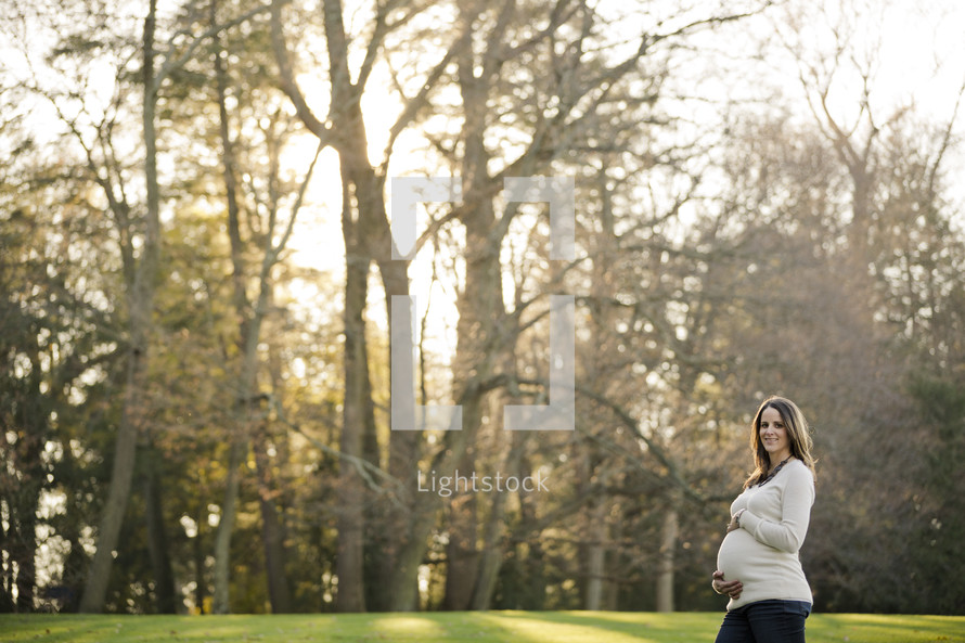woman with her hands on her pregnant belly standing in the grass in front of fall trees