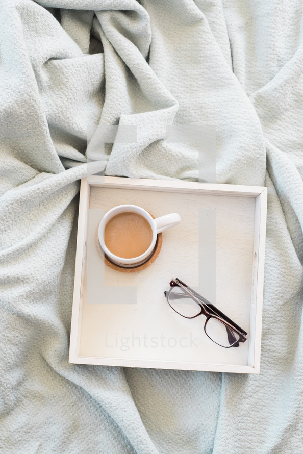 Eyeglasses and a cup of coffee in a square tray on a white cloth.