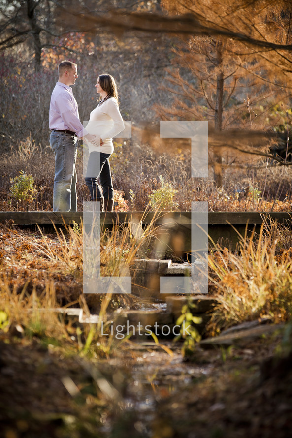 man with his hands on the belly of his pregnant wife standing on a small bridge on a nature trail
