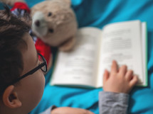Little child, 8 years old boy reading a book at home with his toy teddy bear
