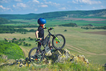 child stands next to his mountain bike on mountains edge and looks at the beautiful scenery