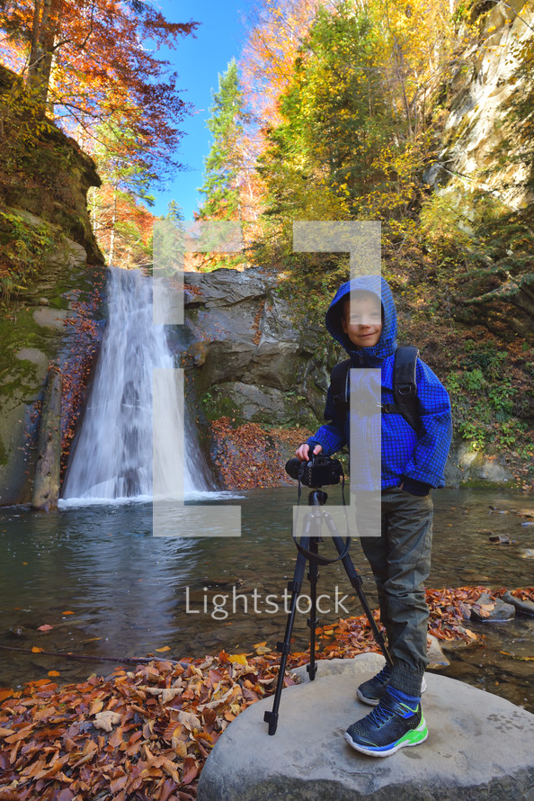 kid near photo camera on tripod with a waterfall in background in autumn