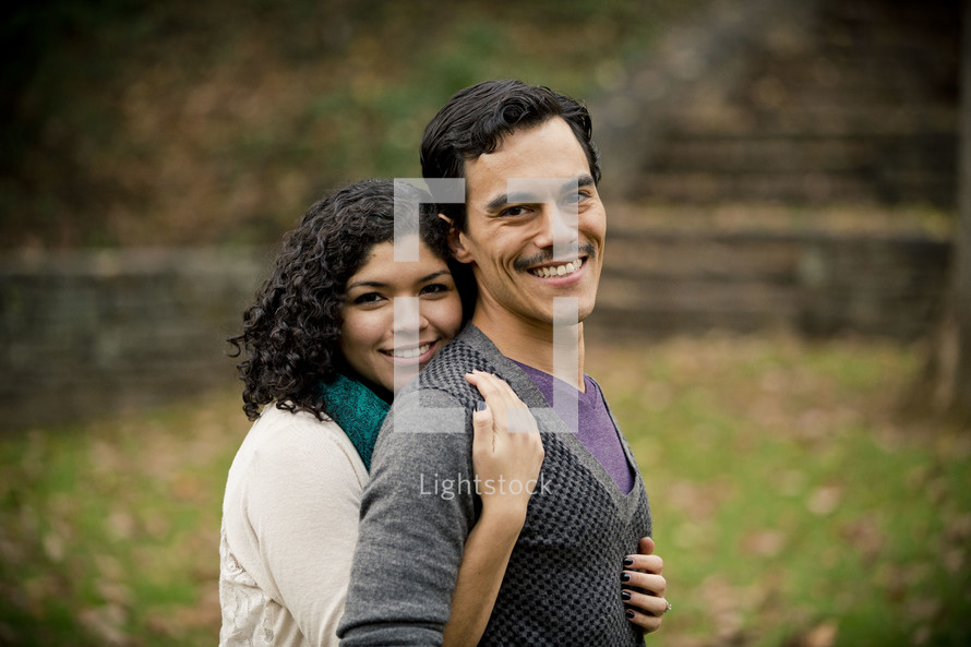 Smiling woman embracing smiling man from behind in park.