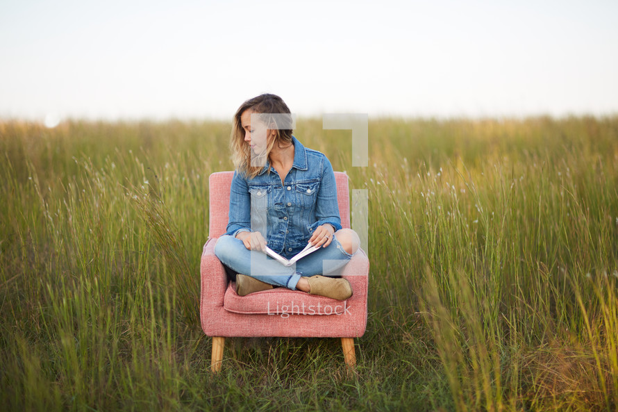 a woman sitting outdoors reading a Bible in a chair in a field of tall grass 