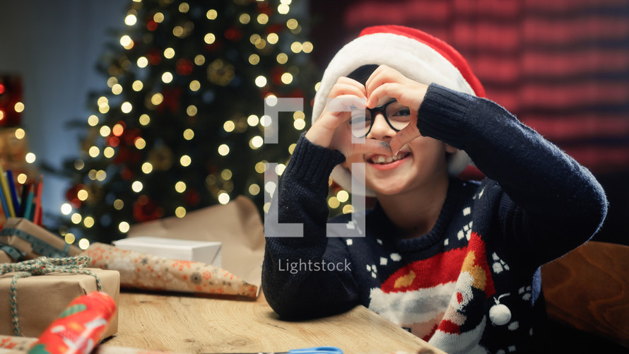 Young Kid under Christmas tree making heart symbol with his hands 
