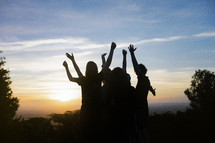 small group of  people standing together with outstretched arms looking out over a valley at sunset