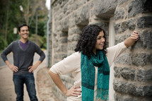 Woman leaning against stone wall with smiling man standing with hands on hips in the background.