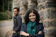 Smiling woman in front of stone wall with smiling man looking at her in the background.