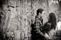 Man and woman leaning against stone wall smiling and embracing.