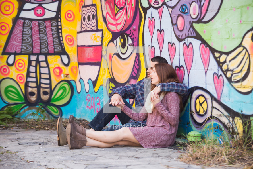 a couple sitting on the ground in front of a wall of street art 