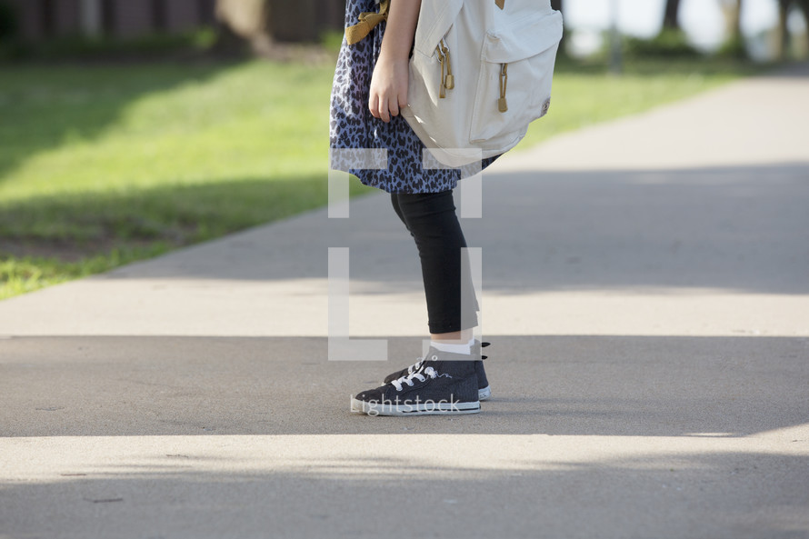 A little girl standing on a sidewalk wearing a backpack.