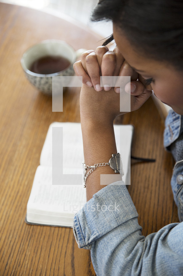 an African American woman sitting at a table reading a Bible and praying
