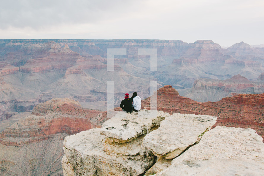people sitting on the edge of a cliff looking out over a canyon 