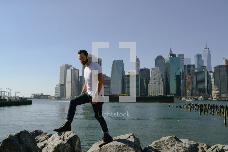 a man standing on rocks along a shore and city buildings in the background 
