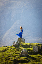 a girl in a dress standing on a rock on a green mountain 
