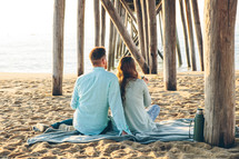 a couple sitting on a blanket under a pier on a beach 