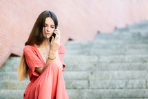 young woman sitting on outdoors stairs wearing red dress talking on a cellphone 