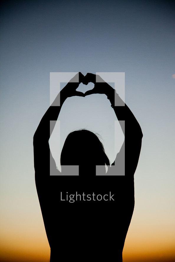 Silhouette of person making a heart shape with their hands with arms extened over head with sunset in background..