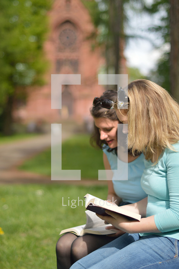 Young women smiling while reading in the bible together outside on a sunny day with a cathedral in the background. 