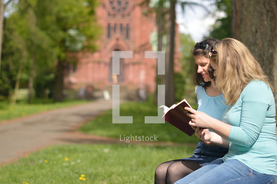 Young women smiling while reading in the bible together outside on a sunny day with a cathedral in the background. 
