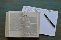 bible studies with a bible open at the book of Judges with notes and pen on a wooden cyan table 