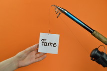 a hand grabbing for a piece of paper with the word FAME on it hanging from a fishing line 