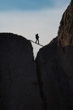silhouette of a person standing on a board stretched between rock peaks 