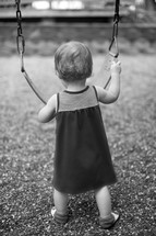 a toddler girl holding a swing in a playground 