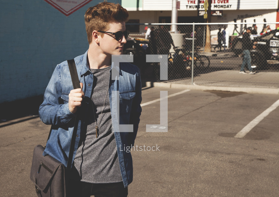 A young man walking through a parking lot carrying a computer bag 