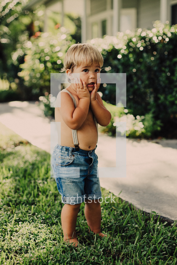 a bare chested toddler boy in suspenders standing in the grass 