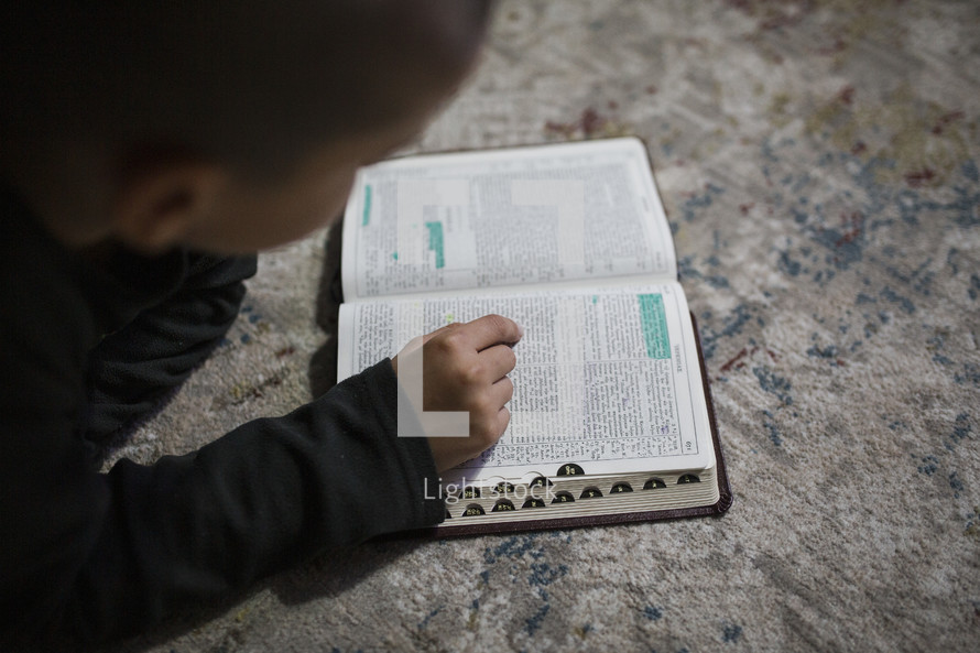 Little boy reading a Bible on the carpet.