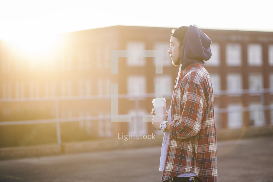 Man in a hoodie standing outside holding a coffee cup at daybreak.