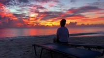 girl sitting on a lounge chair on a beach watching the sunset 