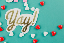 Candy hearts and a paper cutout with the word "yay!" on an aqua background.