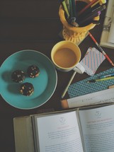cookies on a plate, open Bible, pens in a cup, coffee mug, and journal on a messy desk 
