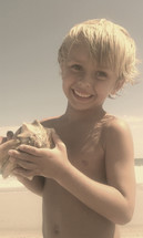 A happy little boy holding a whelk seashell on a beach on a summer day 