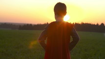 Boy dressed with a Superman cape running in a field, looking into the sunset.