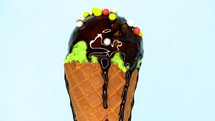 Green pistachio sprinkled ice-cream in waffle cone rotating on blue background and liquid chocolate sauce pouring on it. Delicious isolated sweet dessert in studio