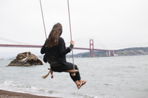 A woman on a swing facing the ocean and the Golden Gate Bridge.