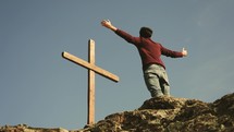 a man standing at the top of a mountain with outstretched arms looking at a cross 