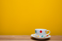 a polka dotted coffee cup and saucer against a yellow wall 