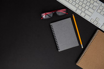 reading glasses, notebook, pencil, and computer keyboard on a desk 