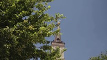 Slow-motion clip looking up at a Church steeple through green tree branches on a beautiful clear blue sky day.