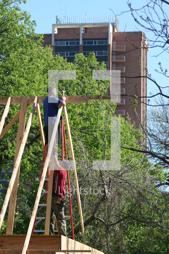 Construction workers putting up roof joists with trees and a tall building in the background.