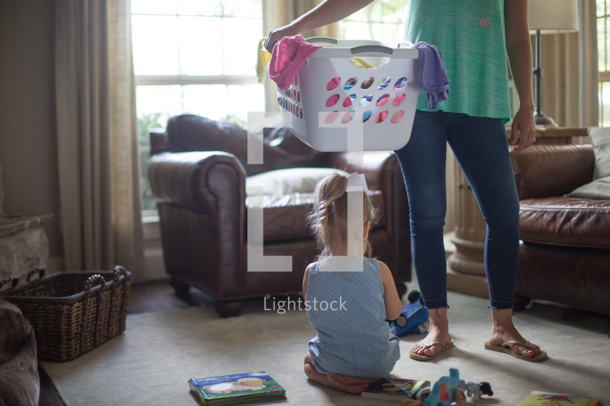 a mother holding a laundry basket with her daughter playing with toys on the floor 