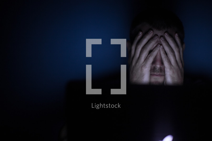  a man covering his eyes looking at a computer screen at night 