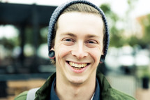 A smiling young man in a winter cap and ear lobe plugs.