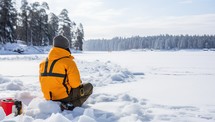 Man in yellow jacket sitting on snow and looking at frozen lake.