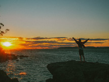 man standing at a shore with raised arms at sunset 